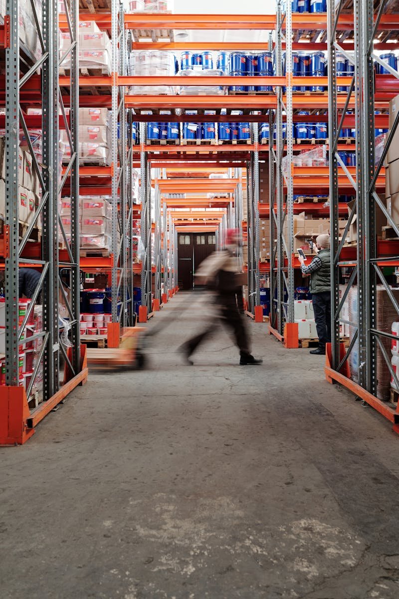 Blurred motion of a man walking in a spacious warehouse aisle between tall shelves.